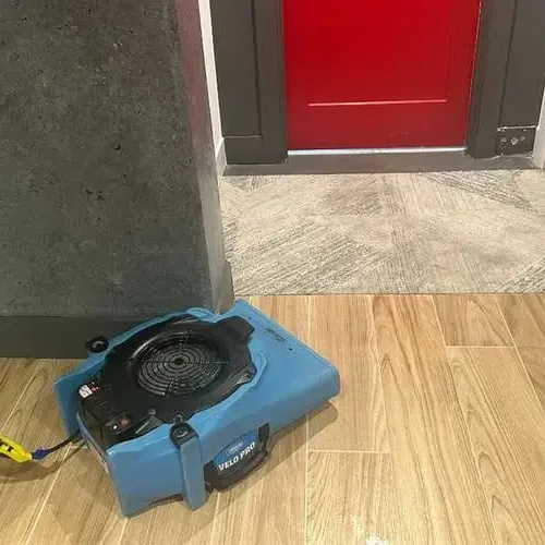 A blue air mover running on a wood look tile floor beside a red door